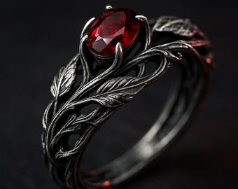 Puede incluir: Un anillo plateado con una gema ovalada de color rojo intenso. El anillo presenta un diseño detallado inspirado en la naturaleza, con hojas y vides. La gema está sujeta por cuatro garras.