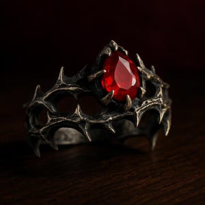 May include: A gothic ring with a dark, textured band featuring sharp, thorn-like details. The ring holds a large, teardrop-shaped, red gemstone. The ring is set against a dark wooden surface.