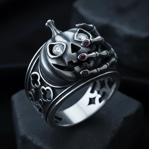Op de afbeelding: Zilverkleurige ring met een gesneden pompoen met diamantachtige ogen en robijnaccenten. De pompoen wordt vastgehouden door skeletachtige handen. De band heeft een decoratief opengewerkt ontwerp. Een uniek Halloween-sieraad.
