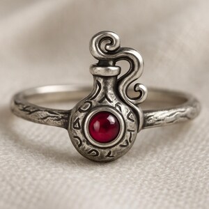 Könnte beinhalten: Ein Silberring mit einem zentralen Motiv in Form einer Zaubertrankflasche, besetzt mit einem tiefroten Cabochon-Edelstein. Der Metallring hat eine texturierte, verwitterte Oberfläche und das Flaschendesign zeigt Gravuren und einen geschwungenen Henkel.