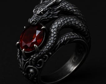 May include: A silver-toned dragon ring with a large, oval-cut, red gemstone. The dragon wraps around the finger, its detailed scales and head framing the gem. The ring has a gothic or fantasy theme.