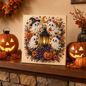 May include: A square diamond painting canvas featuring four white ghosts, a central black lantern, pumpkins, and stars. The artwork is decorated with colorful faceted beads, creating a textured, sparkling Halloween scene with floral accents.