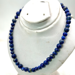 May include: A beaded necklace featuring round, polished lapis lazuli stones in deep shades of blue with subtle natural flecks of gold and white. The beads are uniform in size and strung together to form a continuous circular strand.