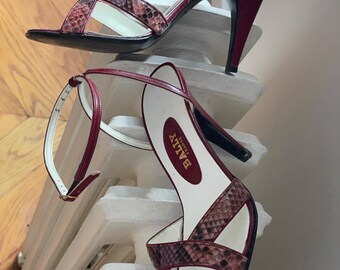 May include: A pair of high-heeled sandals with a snakeskin pattern and burgundy straps. The sandals have a stiletto heel and a white insole with the brand name "BALLY FRANCE" printed on it. The sandals are displayed on a white surface.