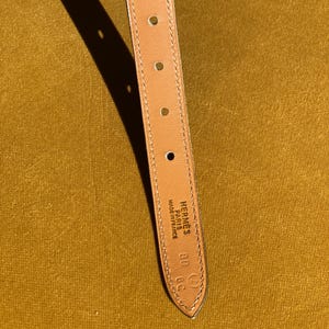 May include: Close-up of a tan leather belt strap with several evenly spaced holes. The strap has a pointed end with the brand name "Hermes Paris Made in France" and size markings. The background is a solid mustard yellow.