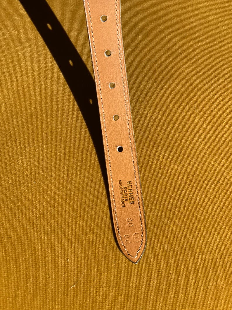 May include: Close-up of a tan leather belt strap with several evenly spaced holes. The strap has a pointed end with the brand name "Hermes Paris Made in France" and size markings. The background is a solid mustard yellow.