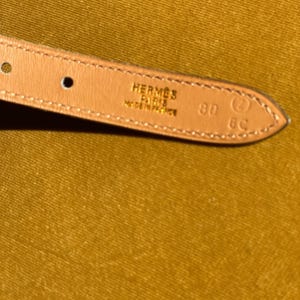 May include: Close-up of a light brown leather belt strap. The strap is stamped with "HERMES PARIS MADE IN FRANCE" and the size "80". The background is a solid mustard yellow.