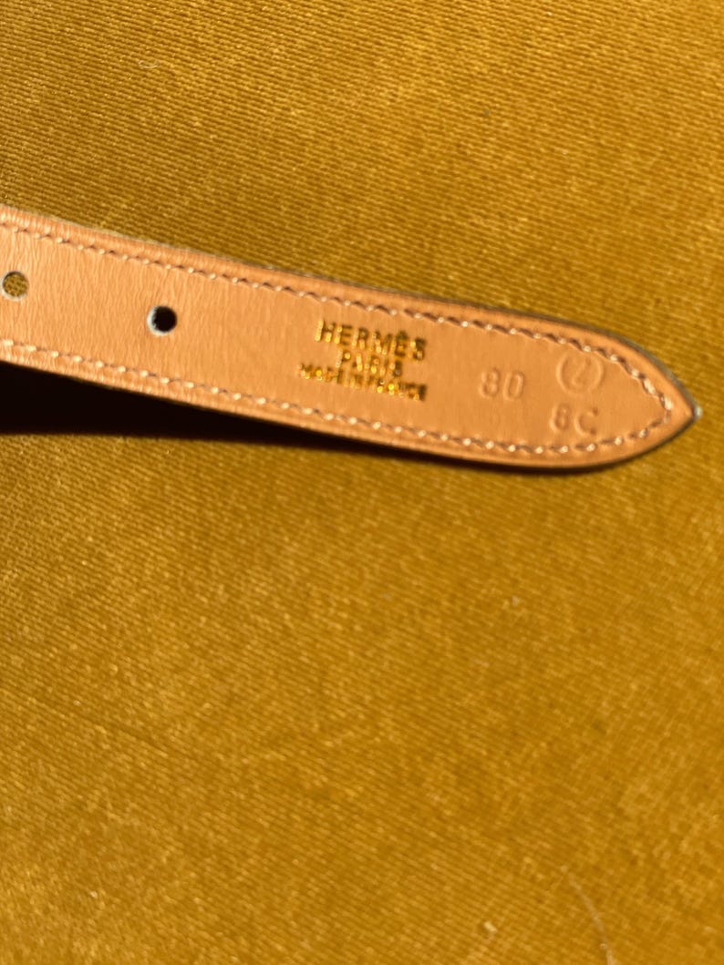 May include: Close-up of a light brown leather belt strap. The strap is stamped with "HERMES PARIS MADE IN FRANCE" and the size "80". The background is a solid mustard yellow.
