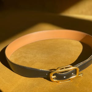 May include: A black leather belt with a gold-colored buckle. The belt's interior is a light tan color. The belt is lying on a yellow surface, with sunlight casting shadows.