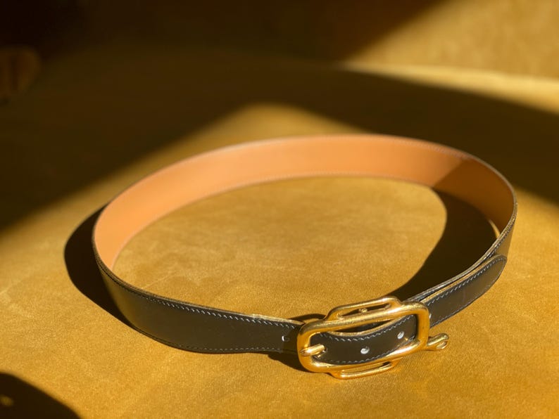 May include: A black leather belt with a gold-colored buckle. The belt's interior is a light tan color. The belt is lying on a yellow surface, with sunlight casting shadows.