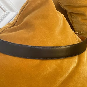 May include: A black leather belt with a gold-colored buckle. The belt is displayed on a yellow velvet surface. The belt is a classic accessory.