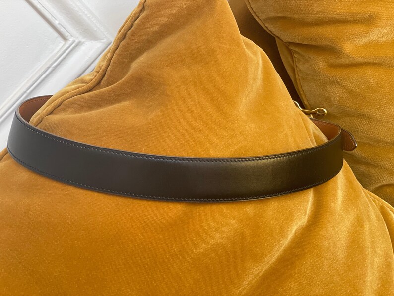 May include: A black leather belt with a gold-colored buckle. The belt is displayed on a yellow velvet surface. The belt is a classic accessory.