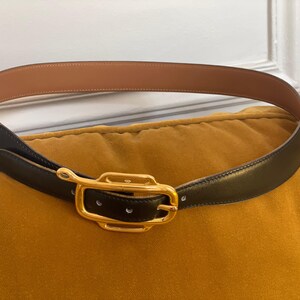 May include: A black leather belt with a gold-colored rectangular buckle. The belt's interior is a light brown color. The belt is resting on a yellow-orange surface.