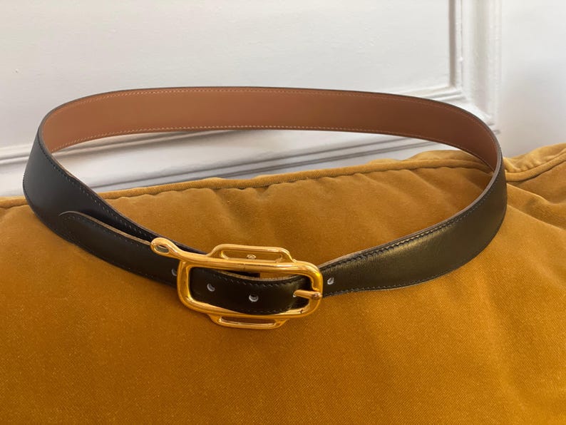 May include: A black leather belt with a gold-colored rectangular buckle. The belt's interior is a light brown color. The belt is resting on a yellow-orange surface.