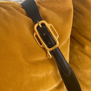 May include: Black leather belt with a gold-colored buckle. The buckle has a unique, ornate design. The belt is shown against a mustard-colored velvet background.