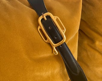 Pode incluir: Cinto de couro preto com fivela dourada. A fivela tem um design ornamental único. O cinto é mostrado sobre um fundo de veludo cor de mostarda.