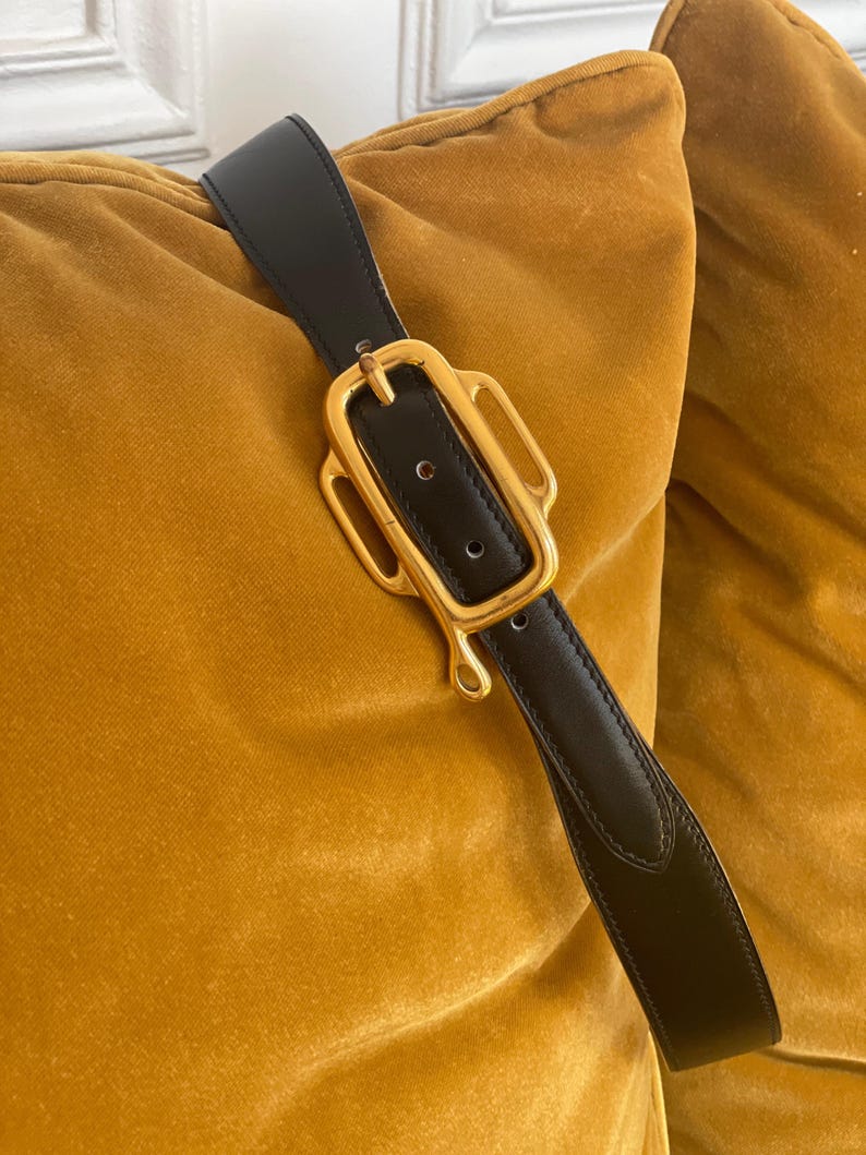 May include: Black leather belt with a gold-colored buckle. The buckle has a unique, ornate design. The belt is shown against a mustard-colored velvet background.