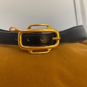 May include: Black leather belt with a gold-colored rectangular buckle. The buckle has a unique design with rounded edges and a small loop on one side. The belt is set against a mustard-colored background.