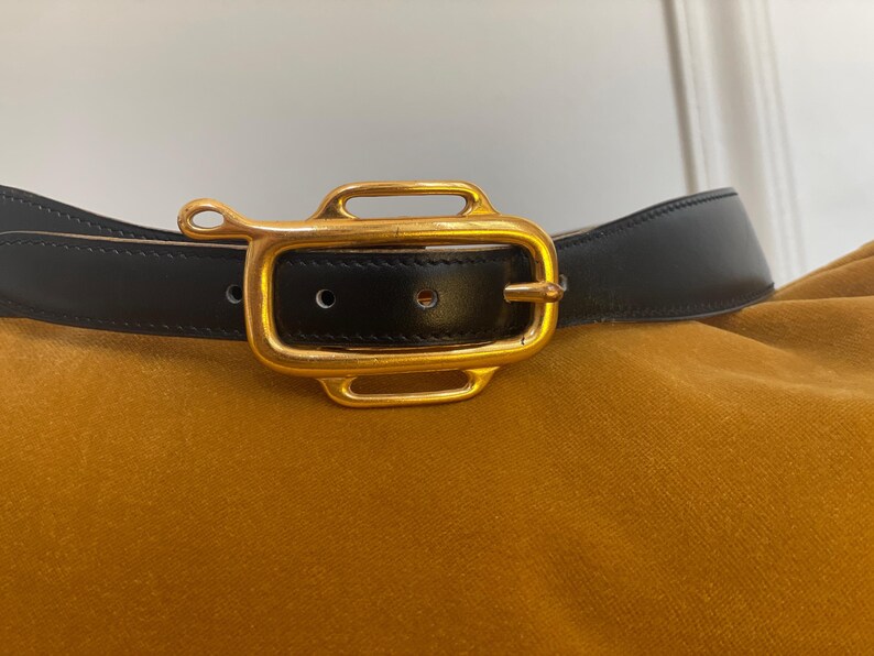 May include: Black leather belt with a gold-colored rectangular buckle. The buckle has a unique design with rounded edges and a small loop on one side. The belt is set against a mustard-colored background.