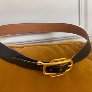 May include: A black leather belt with a gold-colored buckle. The belt has a light brown interior and is resting on a yellow cushion. The buckle is rectangular with a curved design.