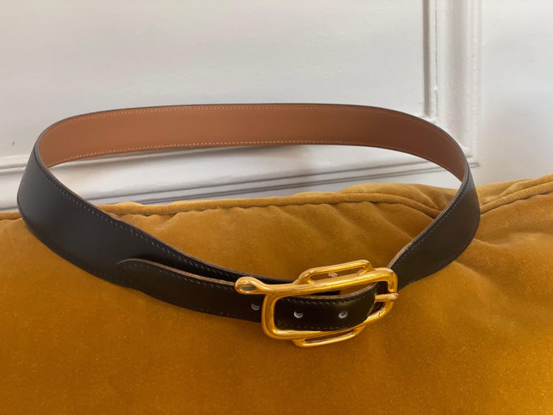 May include: A black leather belt with a gold-colored buckle. The belt has a light brown interior and is resting on a yellow cushion. The buckle is rectangular with a curved design.