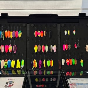 May include: A black tackle box filled with colorful fishing lures. The lures are various shapes and sizes, with a variety of colors including blue, pink, yellow, and green. The lures are arranged neatly in rows.