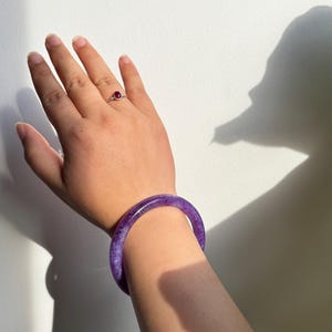 May include: A hand wearing a silver ring with a red gemstone and a purple jade bangle bracelet. The bangle is a solid, translucent purple color. The ring features a small, round, red gemstone. The background is a white wall with shadows.