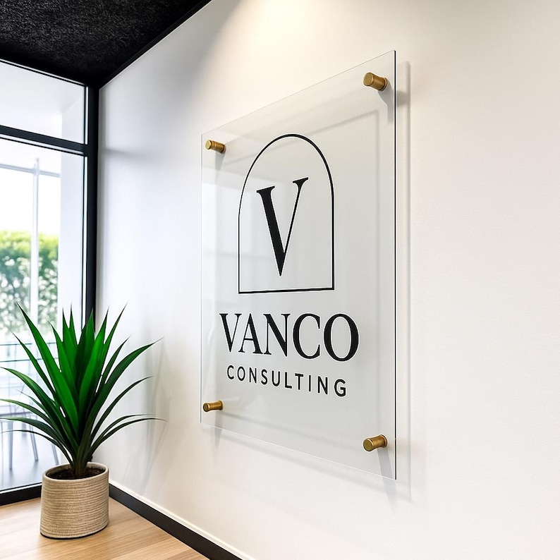 Puede incluir: Un letrero de acr&iacute;lico transparente con el texto "VANCO CONSULTING" en negro. El letrero est&aacute; montado en una pared blanca con separadores dorados. El logotipo presenta una "V" negra dentro de una forma arqueada. Una planta en maceta es visible en la esquina.