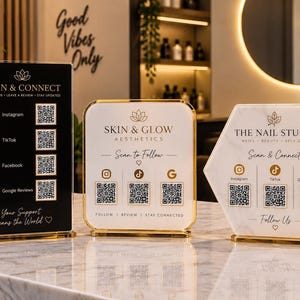 Könnte beinhalten: Drei Acrylschilder mit goldenen Rändern und Ständern. Links ein schwarzes rechteckiges Schild mit vier QR-Codes. In der Mitte ein weißes quadratisches Schild mit abgerundeten Ecken und drei QR-Codes. Rechts ein weißes sechseckiges Schild mit drei QR-Codes.