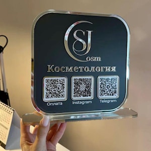 May include: A square black sign with rounded corners and a silver mirrored border. It features the silver logo SJ osm, the text Косметология, and three square QR codes labeled Oплата, Instagram, and Telegram in silver lettering.