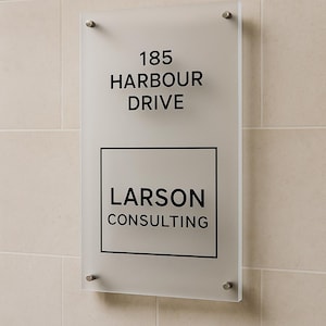 Puede incluir: Un letrero de vidrio esmerilado con la direcci&oacute;n "185 HARBOUR DRIVE" y el nombre "LARSON CONSULTING" en letras negras. El letrero est&aacute; montado en una pared de azulejos beige con cuatro separadores plateados.