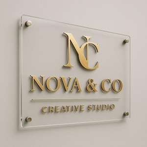 Puede incluir: Letrero de acr&iacute;lico transparente con letras doradas. El letrero dice "NC" encima de "NOVA & CO" y "CREATIVE STUDIO". El letrero est&aacute; montado con cuatro separadores de color plateado en una pared gris claro.