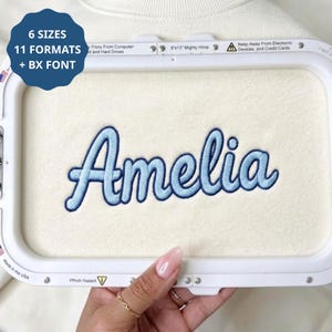 May include: An embroidery hoop containing cream-coloured fabric with the name Amelia stitched in light and dark blue thread. A blue circular graphic in the corner displays the text 6 SIZES, 11 FORMATS, + BX FONT. The hoop is labelled 8x13 Mighty Hoop.
