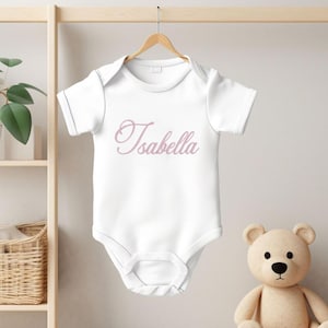 May include: A white short-sleeved baby bodysuit featuring the name Isabella printed in a light pink cursive script across the chest. The garment has a round neckline and a snap-button closure at the bottom, hanging on a wooden hanger.