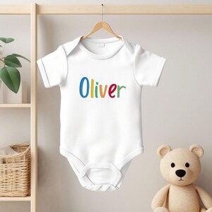 May include: White baby onesie with the name "Oliver" embroidered in colorful letters. The onesie is hanging on a wooden hanger against a light background. A teddy bear is in the background.