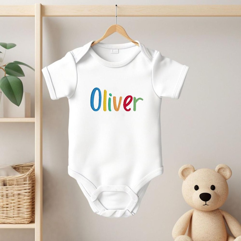 May include: White baby onesie with the name "Oliver" embroidered in colorful letters. The onesie is hanging on a wooden hanger against a light background. A teddy bear is in the background.