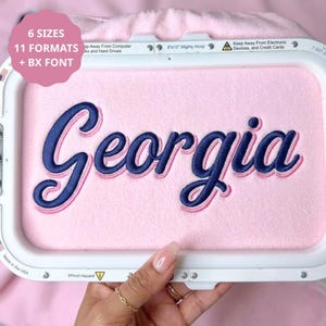 May include: An embroidery hoop containing light pink fabric with the name Georgia stitched in dark blue thread with a light pink outline. The hoop frame includes text stating 6 SIZES, 11 FORMATS, and BX FONT, along with safety warnings.