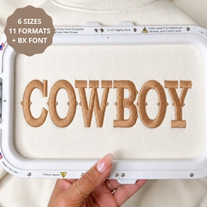 May include: A white fabric surface held in a white embroidery hoop featuring the brown embroidered text COWBOY. A brown circular graphic in the top left corner displays the text 6 SIZES 11 FORMATS + BX FONT. The hoop frame includes safety text.