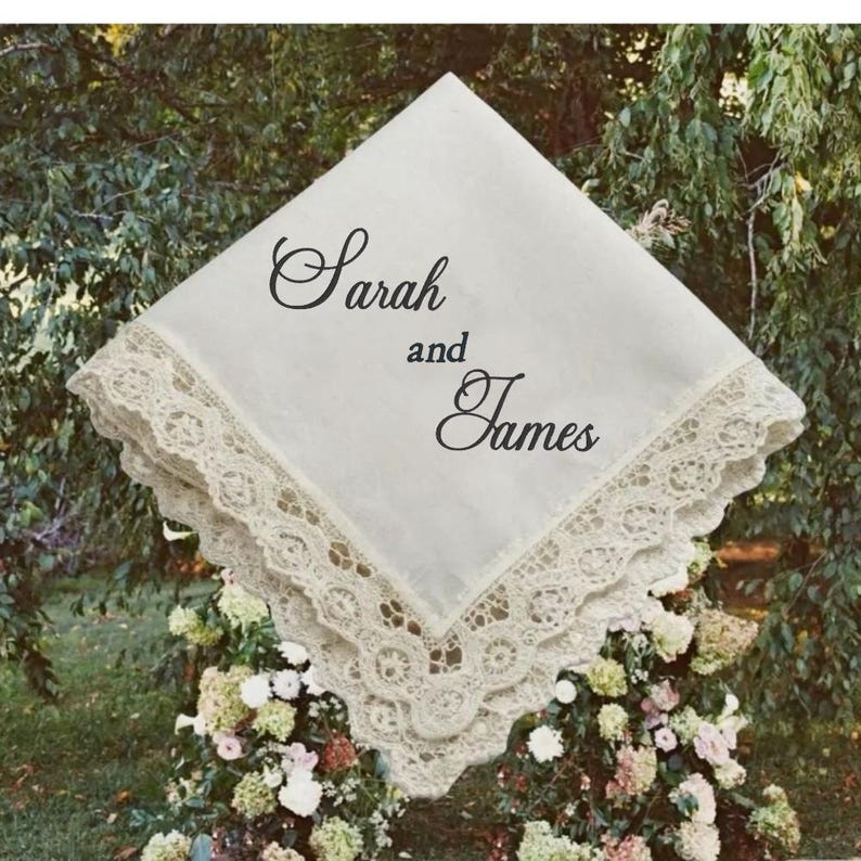 May include: A square white fabric handkerchief featuring a wide, intricate cream-colored lace border. The center of the fabric is printed with the black text Sarah and James in a decorative script font, arranged in three distinct lines.