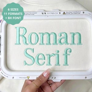 May include: An embroidery hoop containing white fabric with the mint green embroidered text 'Roman Serif'. The hoop frame includes the text '8x13 Mighty Hoop' and a label stating '6 SIZES 11 FORMATS + BX FONT'. The frame is white plastic.