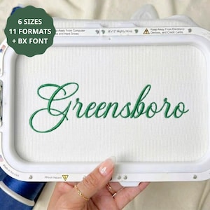 Könnte beinhalten: Ein weißer Stickrahmen mit weißem Stoff, auf dem das Wort Greensboro in grüner Schreibschrift gestickt ist. Der Rahmen enthält den Text 6 SIZES, 11 FORMATS + BX FONT, 8x13 Mighty Hoop sowie Warnhinweise zu elektronischen Geräten.
