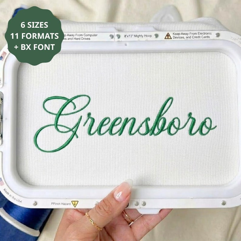 May include: A white embroidery hoop frame containing white fabric with the green embroidered word Greensboro in a cursive script. The frame features text stating 6 SIZES, 11 FORMATS + BX FONT, 8x13 Mighty Hoop, and warnings about electronic devices.