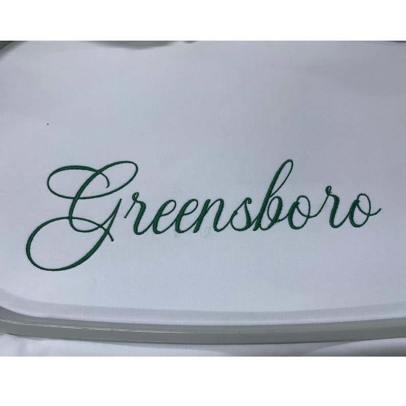 May include: A white fabric surface featuring the word Greensboro embroidered in a dark green cursive script font. The embroidery is centered on the material, which is held taut within a circular embroidery hoop frame visible at the edges.