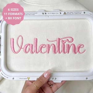 Könnte beinhalten: Ein Stickrahmen mit weißem Stoff und dem in Rosa gestickten Wort Valentine, das ein kleines Herz über dem Buchstaben i aufweist. Der Rahmen enthält den Text 6 SIZES, 11 FORMATS + BX FONT, 8x13 Mighty Hoop und Keep Away From Electronic Devices.