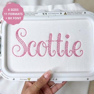 May include: An embroidery hoop containing white fabric with the name Scottie stitched in pink thread. The hoop frame includes text labels indicating 6 sizes, 11 formats, and BX font, along with safety warnings about electronic devices and hard drives.