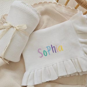 May include: A white knitted baby blanket with the name "Sophia" embroidered in pastel colors. The blanket has a ruffled edge and is folded with a matching ribbon. A second blanket is visible in the background.