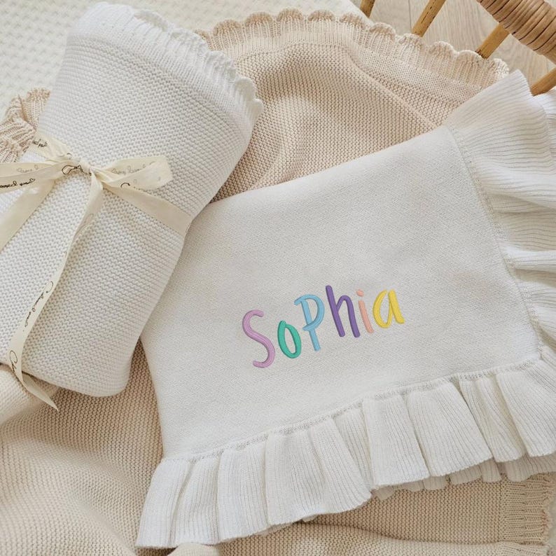 May include: A white knitted baby blanket with the name "Sophia" embroidered in pastel colors. The blanket has a ruffled edge and is folded with a matching ribbon. A second blanket is visible in the background.