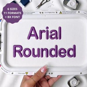 Peut inclure: Un tambour à broder blanc contenant du tissu blanc avec le texte brodé violet Arial Rounded. Un graphique circulaire violet dans le coin supérieur gauche affiche le texte 6 SIZES 11 FORMATS + BX FONT. Le cadre du tambour est en plastique blanc.
