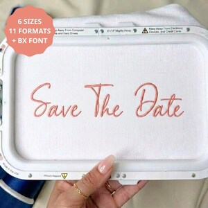 May include: A white embroidery hoop frame containing white fabric with the words Save The Date stitched in coral pink thread. The frame features text indicating 6 sizes, 11 formats, and BX font, along with an 8x13 inch Mighty Hoop label.