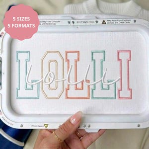 May include: An embroidery hoop containing white fabric with the word LOLLI stitched in a multi-colored varsity font. The letters are outlined in light blue, peach, and pink, with a white cursive script overlay. The frame is labeled 8x13 inches.